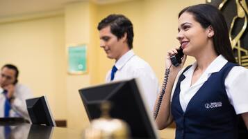A hotel reception area with a receptionist and a guest. There are desks with computers and a clock on the wall.