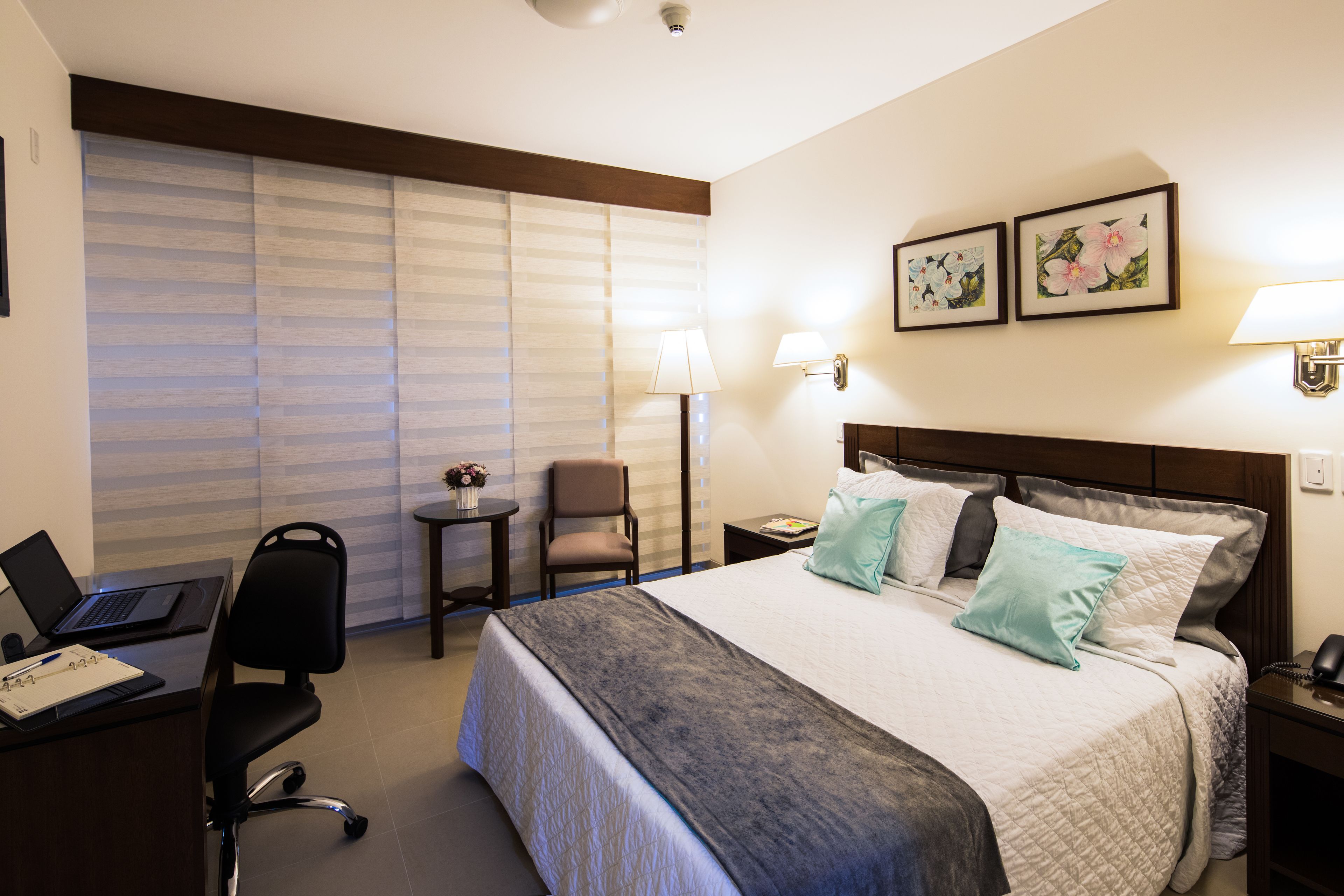 Une chambre d’hôtel comprenant un lit, un bureau avec un ordinateur portable, une chaise, une petite table avec une lampe et deux cadres photos fixés au mur.
