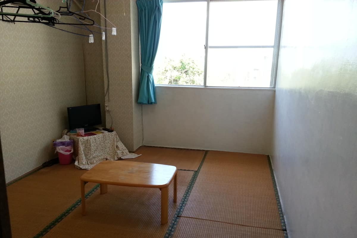 Ein kleiner Raum mit Tatami-Boden, einem Holztisch, einem Fernseher auf einem Standfuß und einem Fenster mit Vorhang.