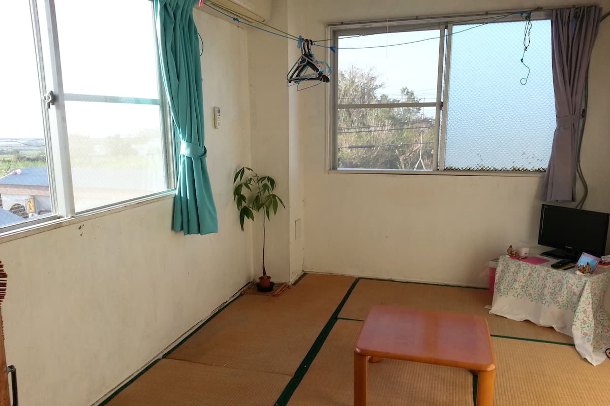 Ein Zimmer mit Tatami-Böden, einer Holzbank, einem Blumentopf, einem Fernseher auf einem Tisch und einem Fenster mit Blick auf Bäume.