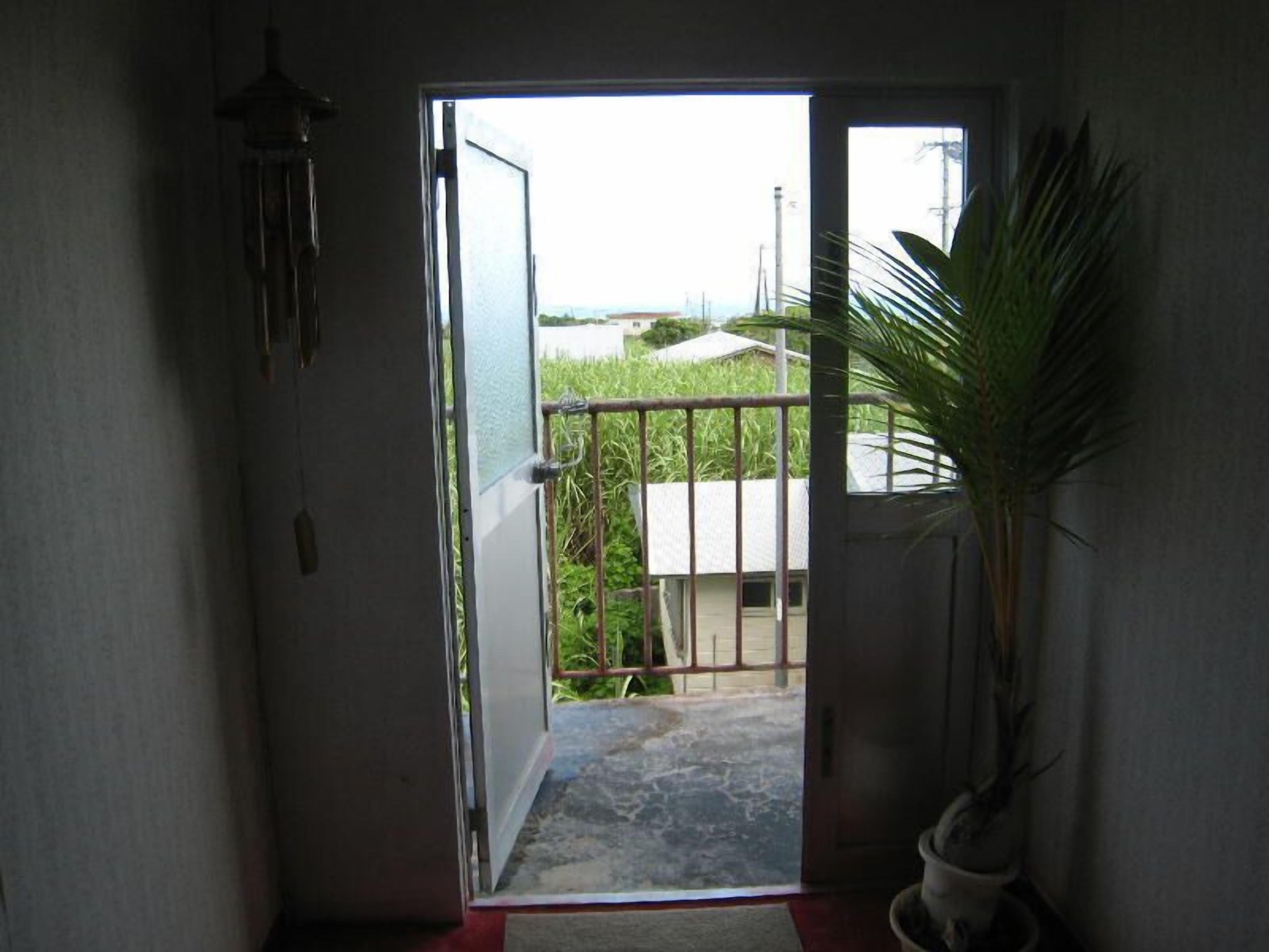 An open door leading to a balcony with a metal gate, a potted plant, and a view of a building and greenery outside.