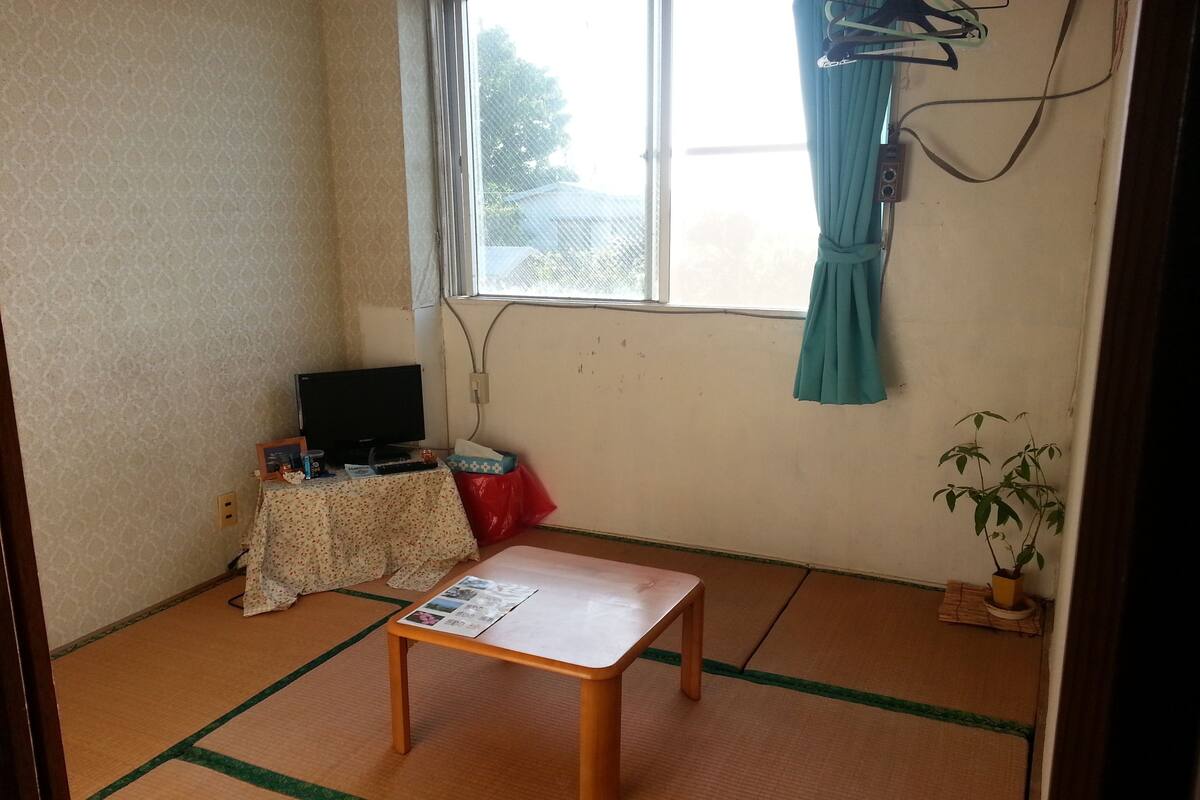 Ein Zimmer mit Tatami-Boden, einem kleinen Holztisch, einem Fernseher auf einem Standfuß, einer Topfpflanze und einem Fenster mit Vorhang.