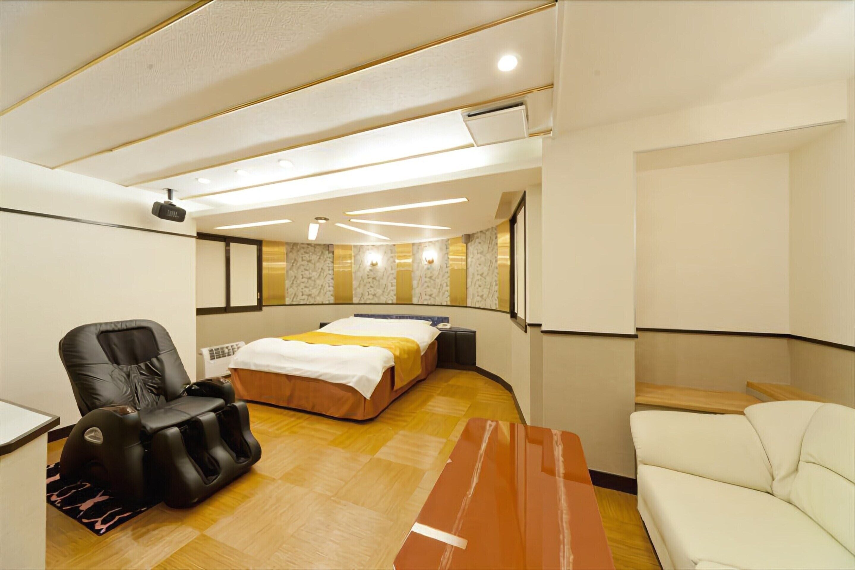 A modern hotel room with a massage chair, a bed with a white and yellow bedspread, a wooden floor, and a white sofa.