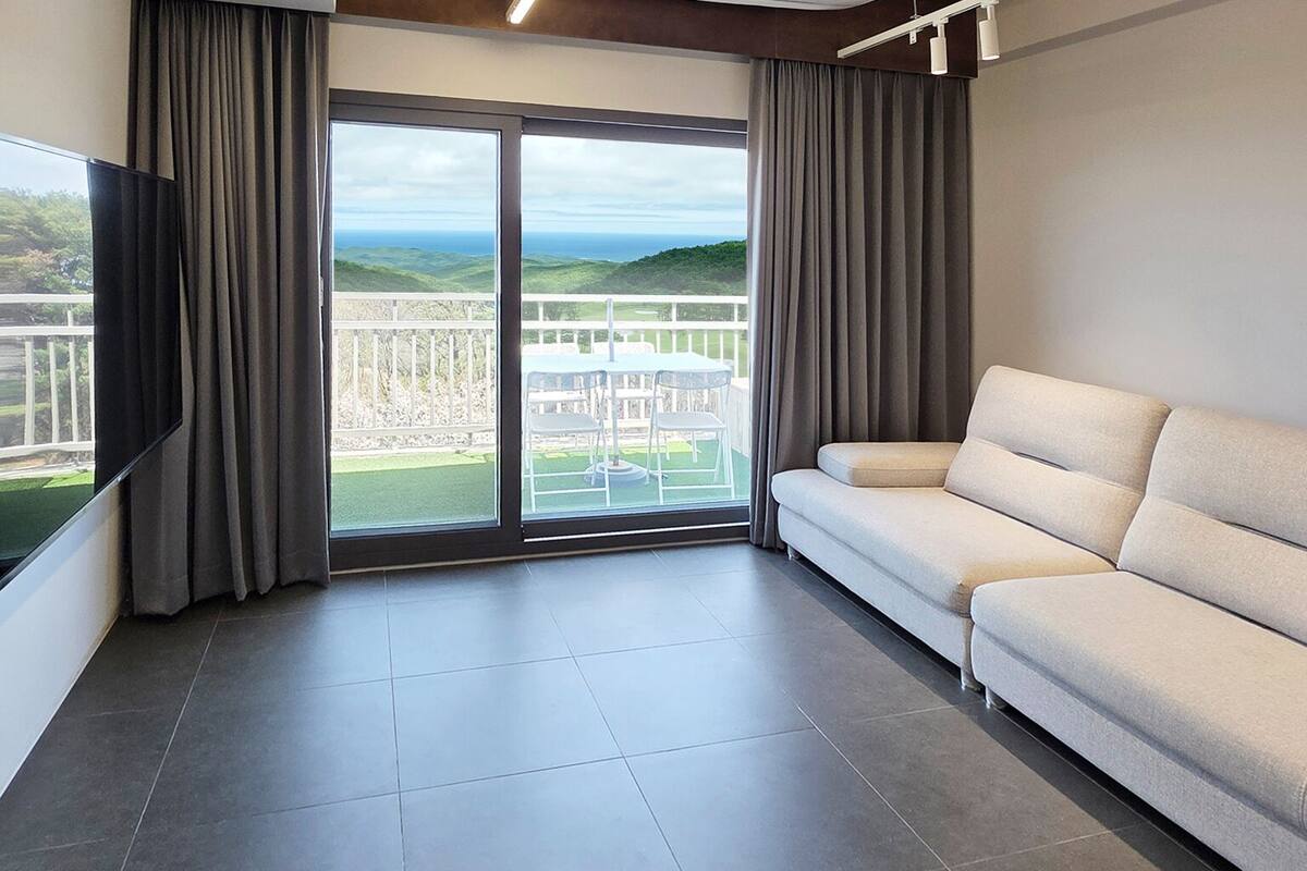A modern living room with a sofa, a flat-screen TV, and a sliding glass door leading to a balcony with a view of the landscape.