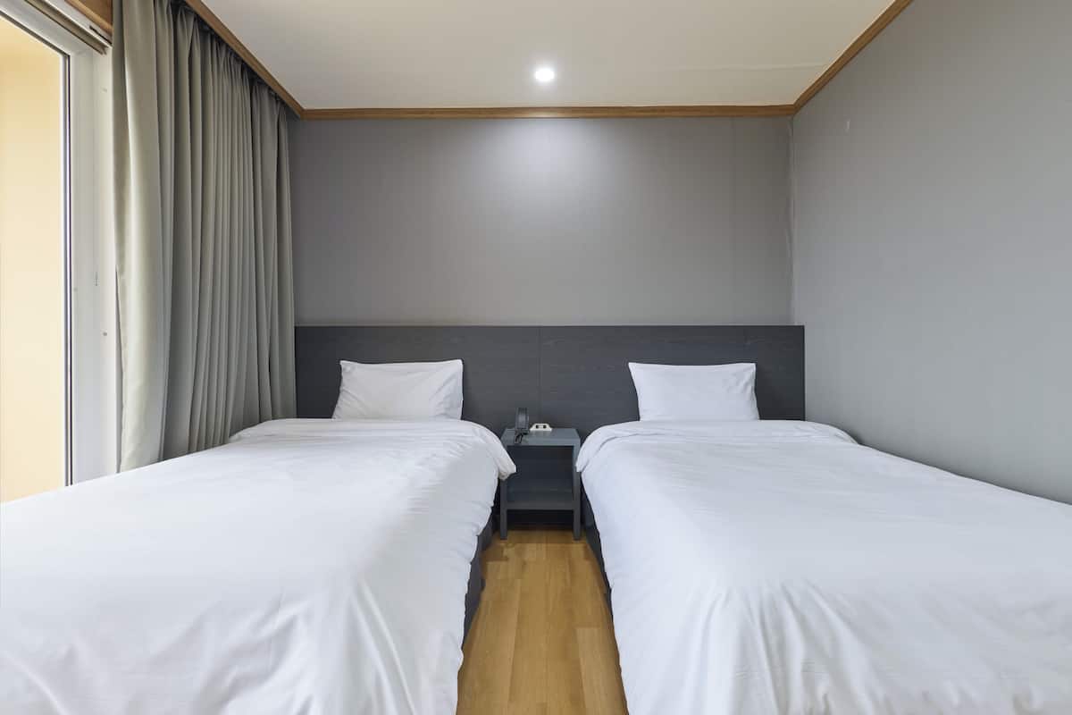 A hotel room with two single beds, a wooden floor, a gray headboard, a bedside table with a phone, and a window with curtains.