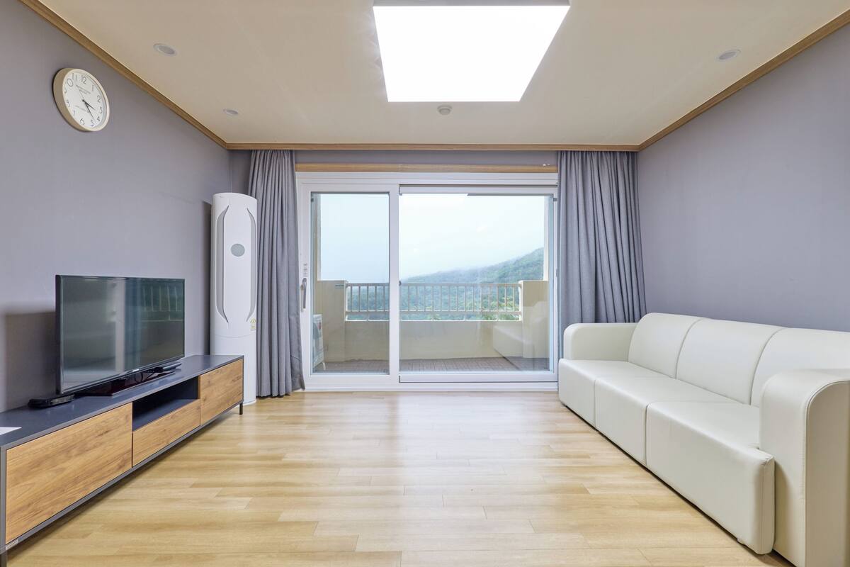 A modern living room with a flat-screen TV, wooden entertainment unit, white sofa, and a sliding glass door leading to a balcony with a view.