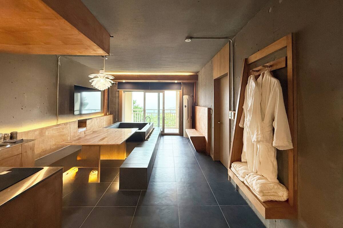 A modern bathroom with a large sink, bench, and a robe hanging on a wooden rack.