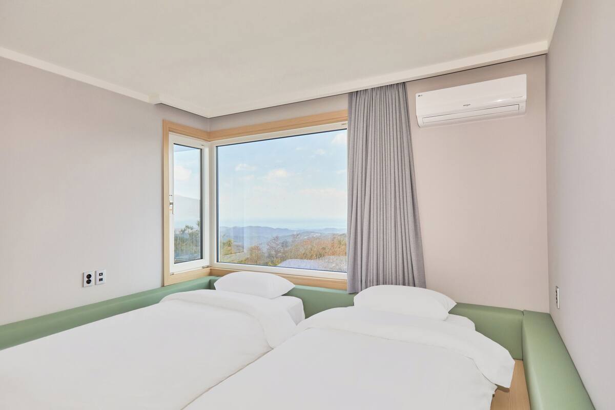A hotel room with two beds, a large window with a view of the outdoors, and an air conditioning unit on the wall.