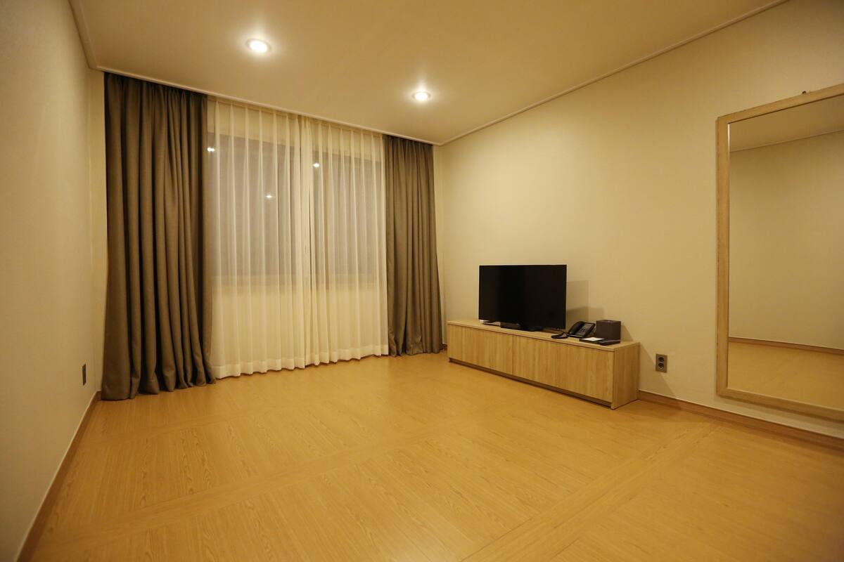 A room with a large mirror, a flat-screen TV on a wooden stand, and a window with sheer and opaque curtains.