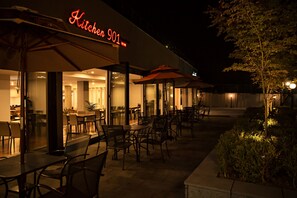 Un espace extérieur aménagé pour les repas, avec des tables et des chaises, un panneau lumineux au néon indiquant « Kitchen 901 », et une entrée bien éclairée du restaurant.