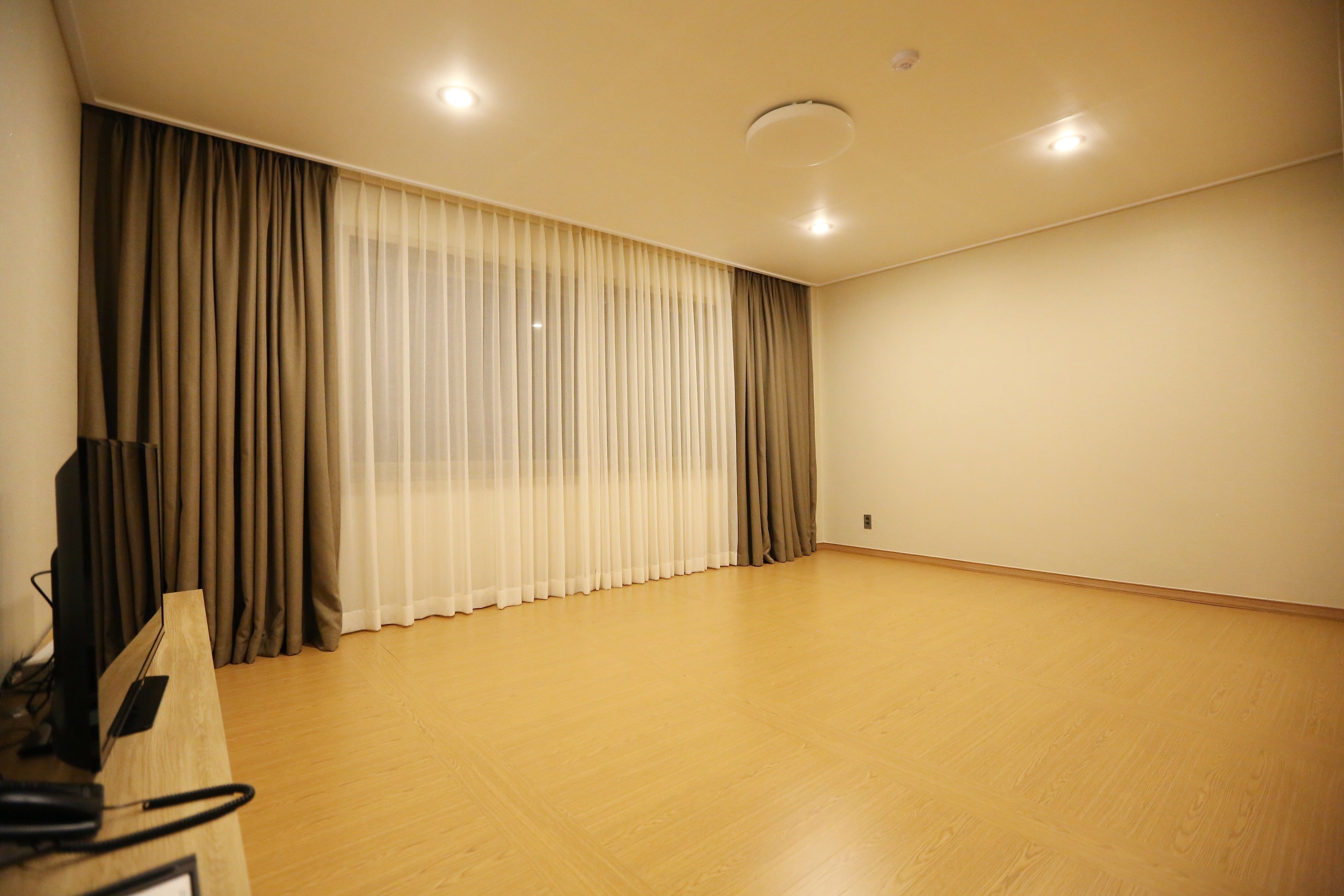Una habitación con piso de madera, un ventanal con cortinas traslúcidas y gruesas, y un televisor de pantalla plana sobre un tocador de madera.