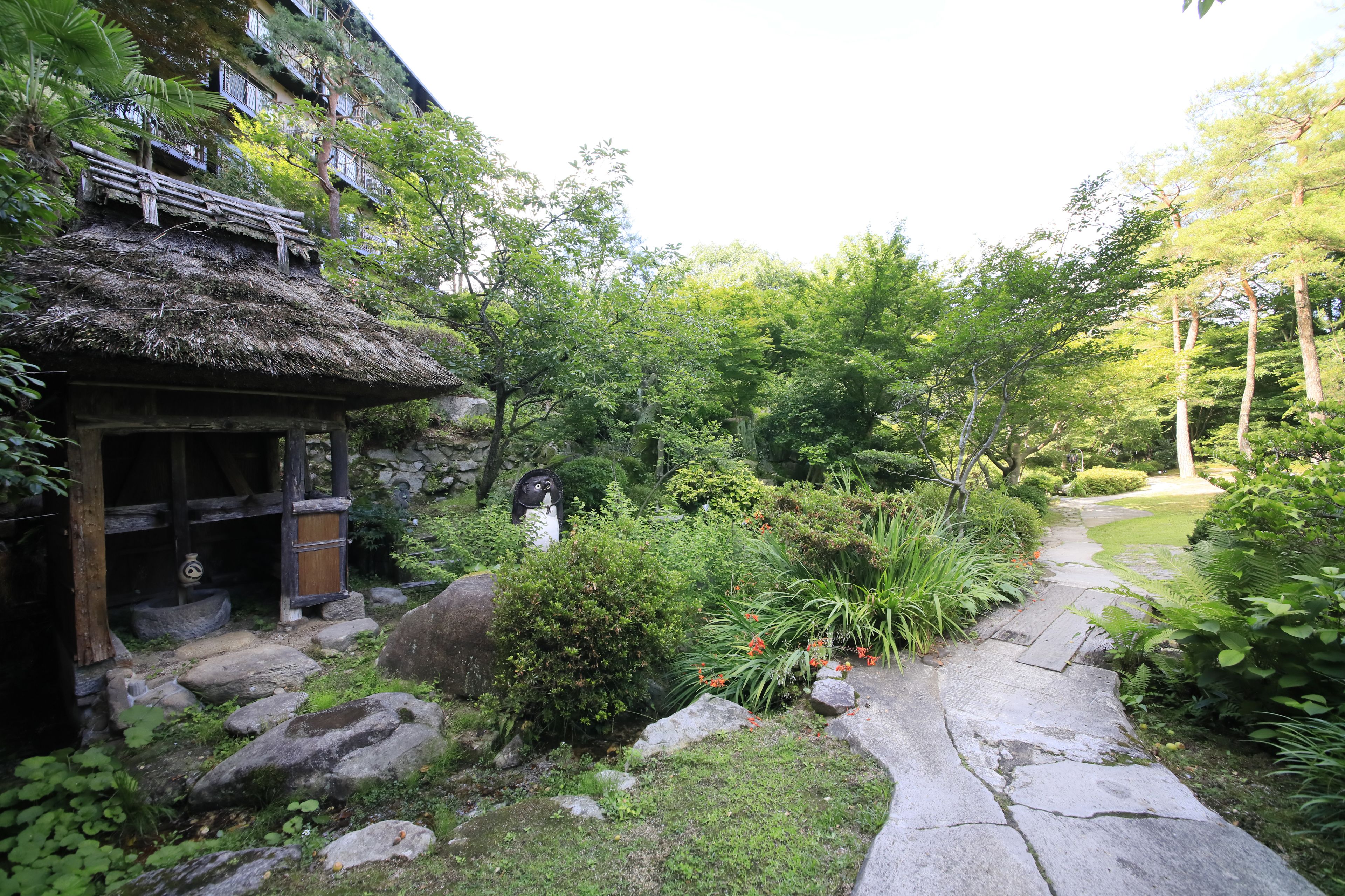Un jardín japonés tradicional con un sendero de piedra, construcciones de madera y abundante vegetación.