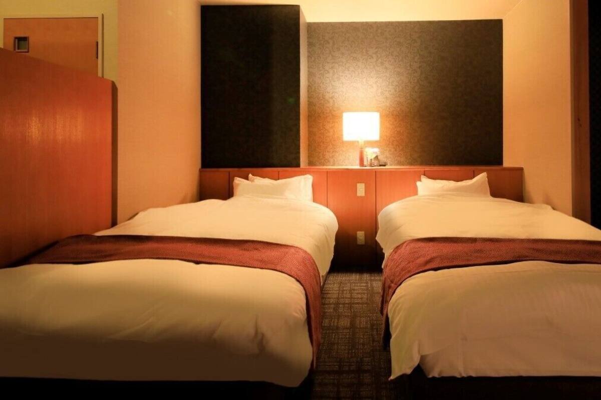 A hotel room with two beds, a nightstand with a lamp, and a large dark wall above the beds.