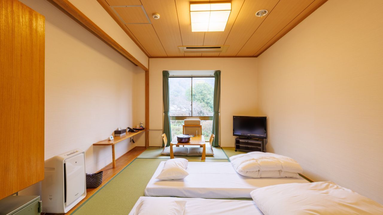 A hotel room with two beds, a TV, a window with curtains, a small table with a bowl, and a radiator.