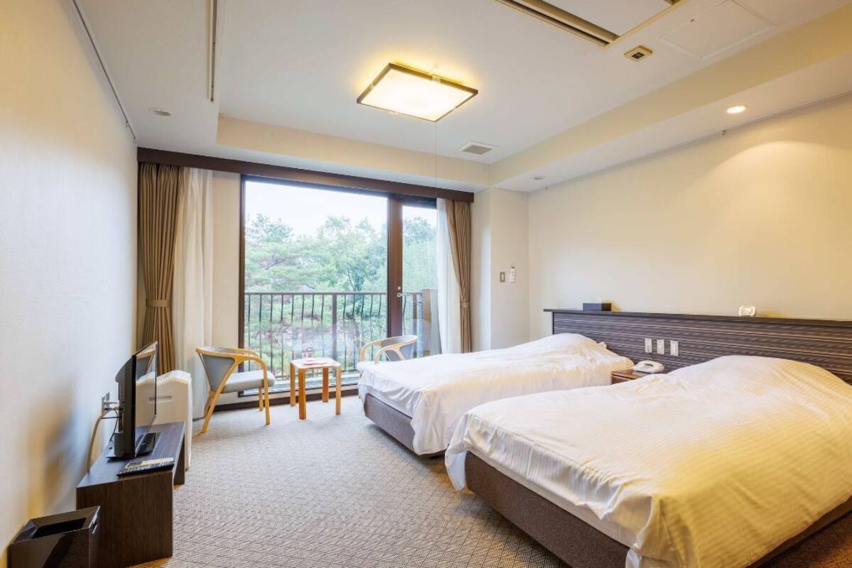 A hotel room with two beds, a desk with a computer, a chair, and a balcony with a view.