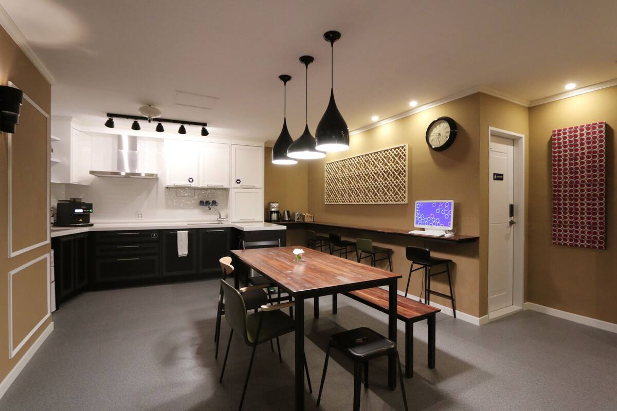 A modern kitchen with white cabinets, a wooden dining table, and black chairs. There is a clock on the wall and a computer on a counter.