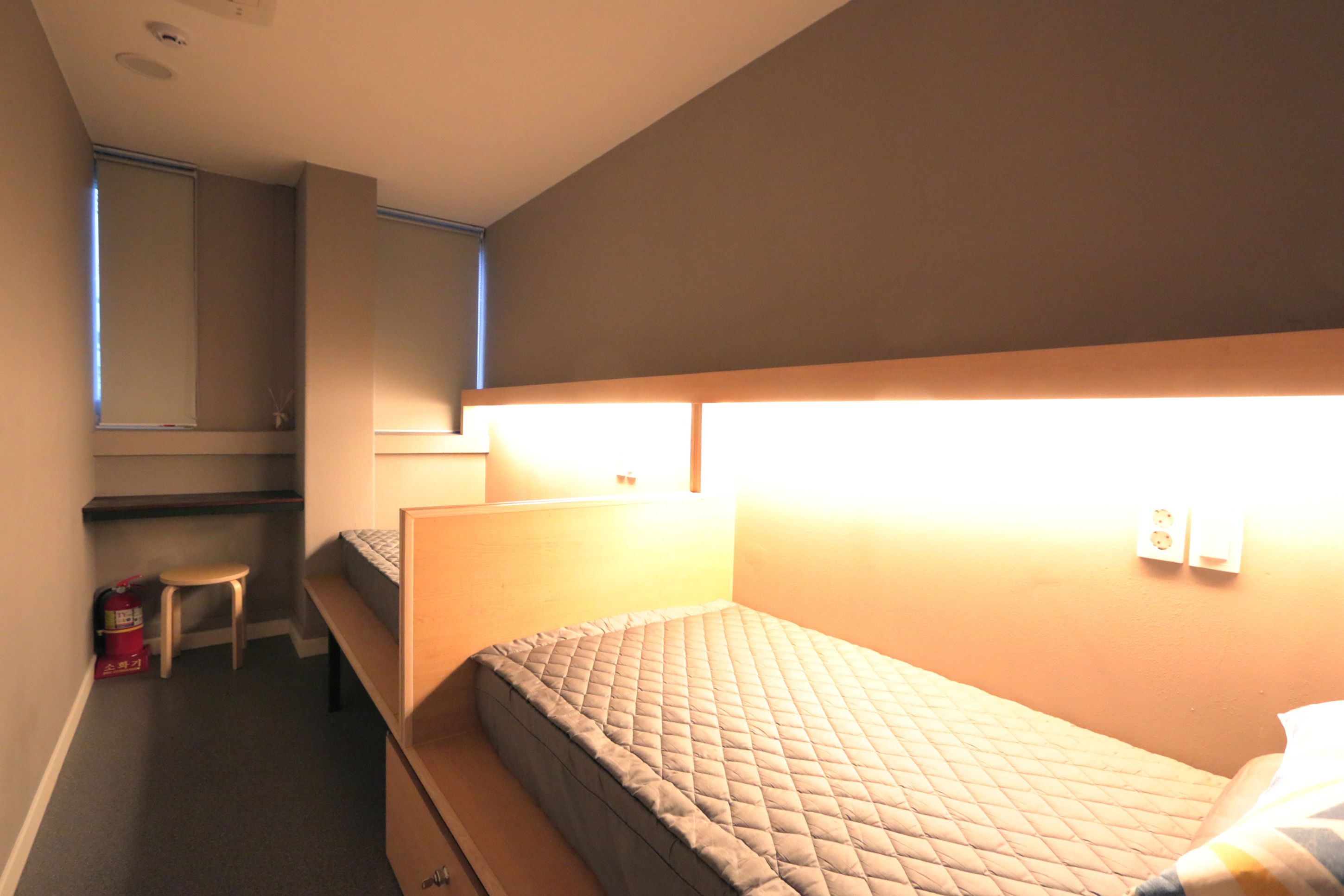 A small, dimly lit room with a single bed, a small stool, and a desk with a chair.