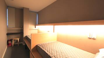 A small, dimly lit room with a single bed, a small stool, and a desk with a chair.
