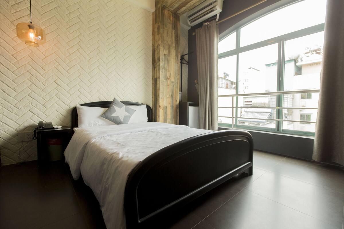 A modern bedroom with a bed, a nightstand, a hanging light, and a large window with a view of buildings.