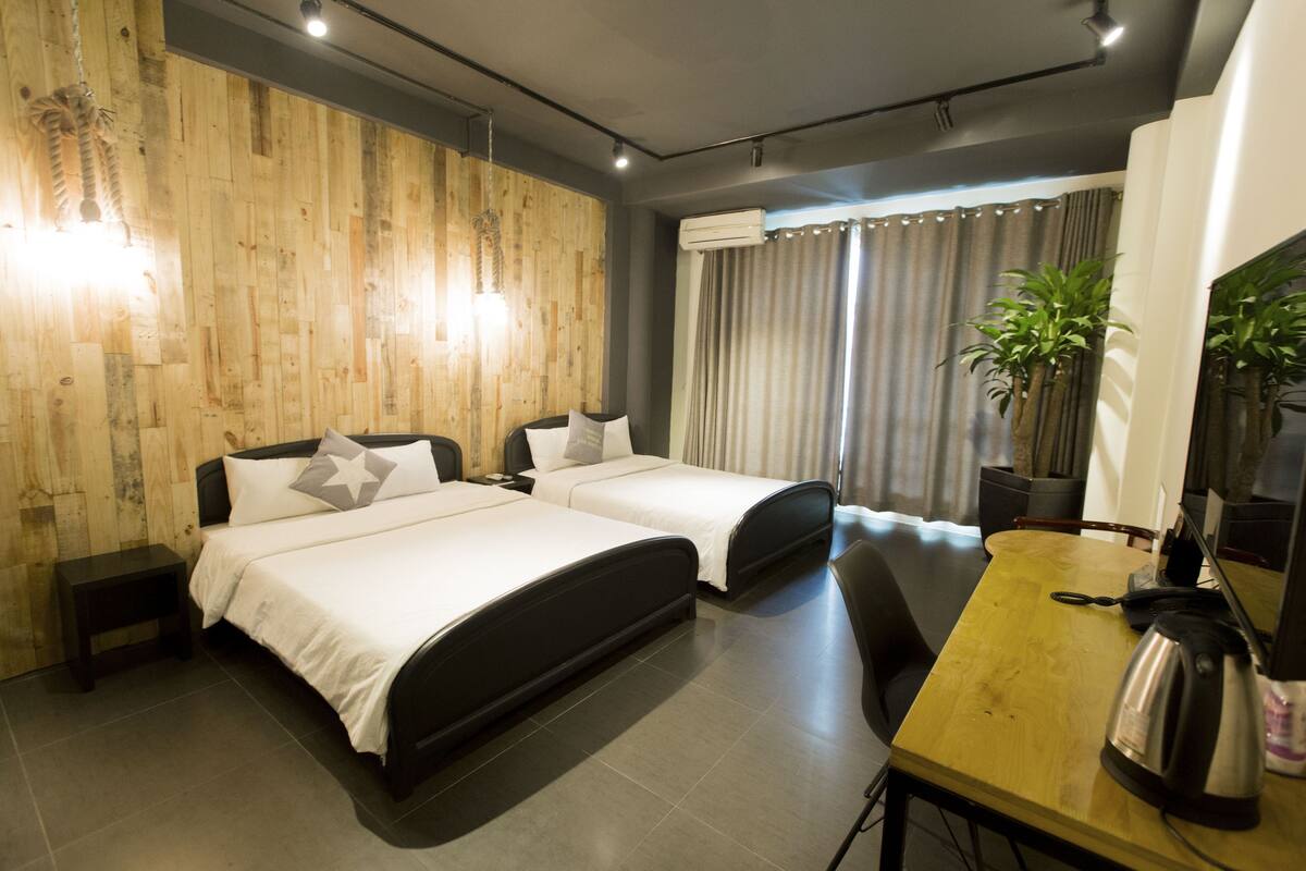 A hotel room with two beds, a wooden wall, a desk with a kettle, and a potted plant.