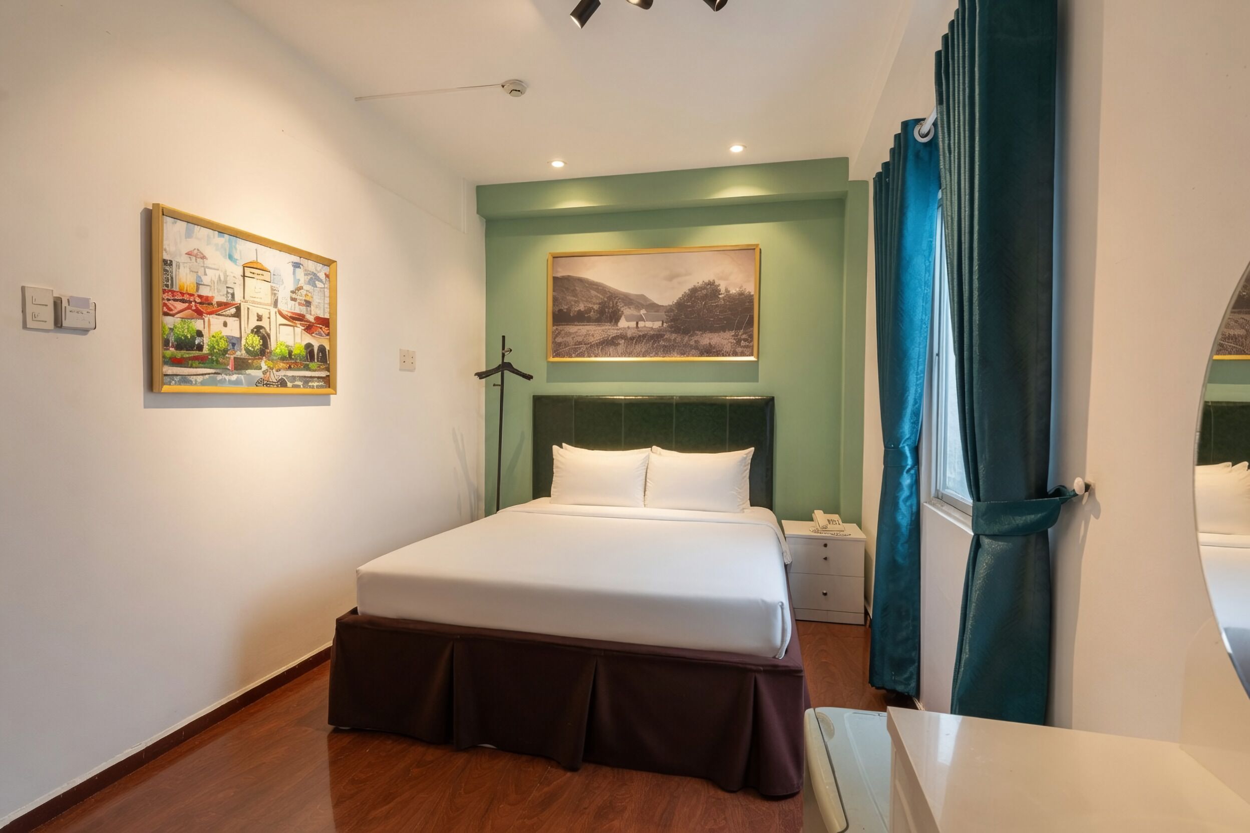 A hotel room with a bed, a nightstand, a lamp, a painting on the wall, and a window with curtains.