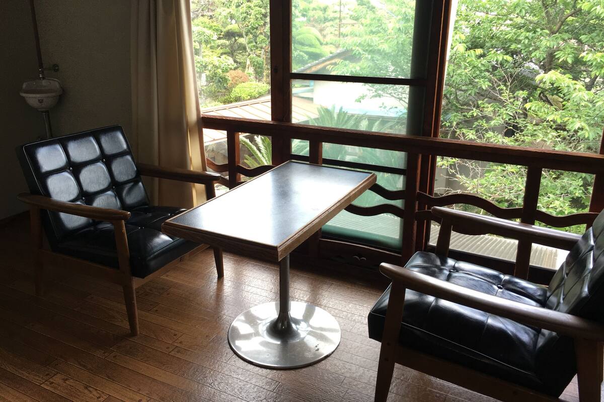 A room with a wooden floor, two black leather chairs, a small table, and a large window with a view of greenery.
