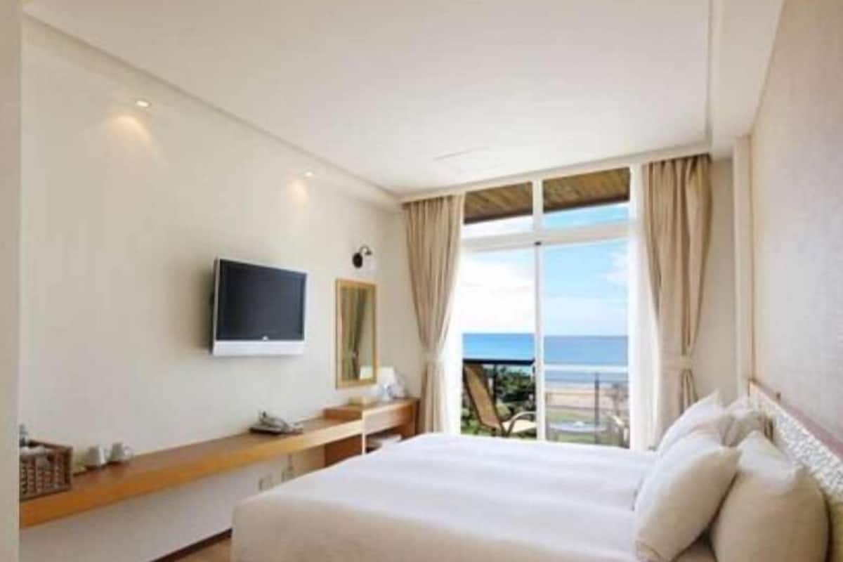A hotel room with a bed, a TV mounted on the wall, a desk with a lamp, and a view of the sea through large windows.