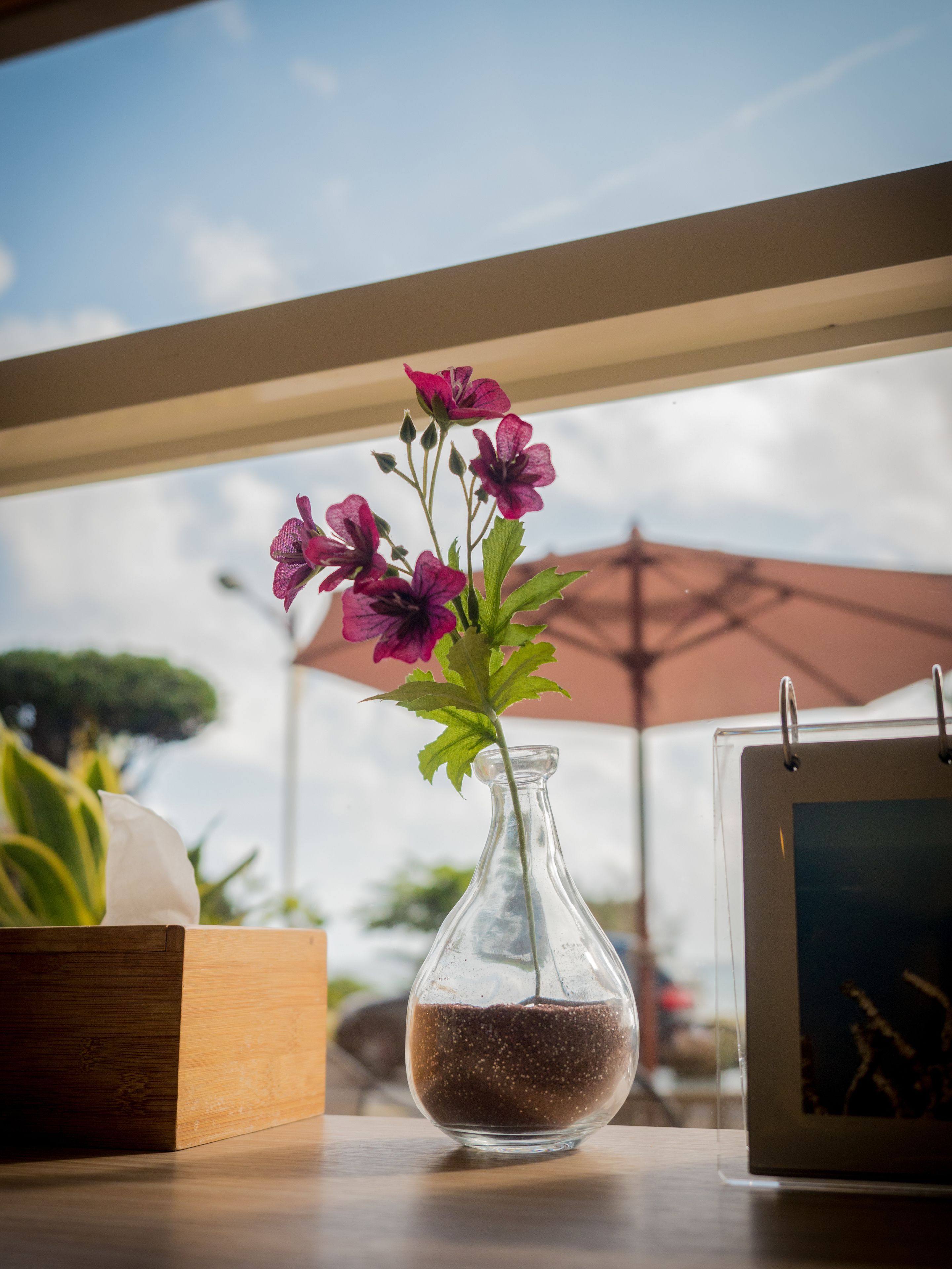 Un jarrón de vidrio transparente con flores moradas sobre una mesa de madera, una tableta y una caja de madera.