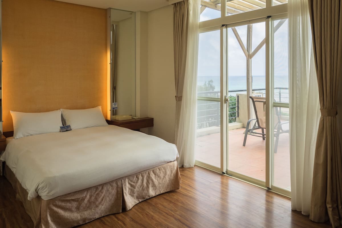 A hotel room with a large bed, a wooden headboard, a nightstand, a mirror, and a balcony with a chair.