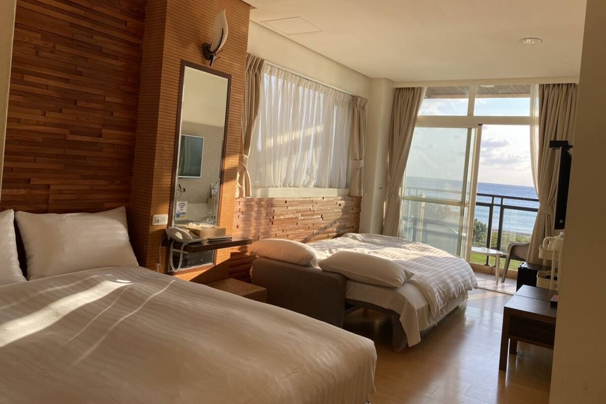 A hotel room with two beds, a large window with a view of the sea, a wooden accent wall, and a desk with a chair.