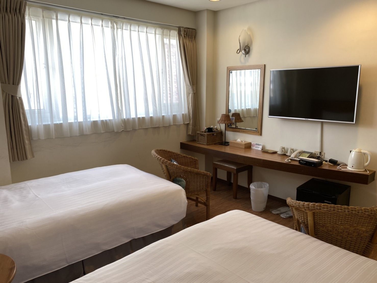 A hotel room with two single beds, a desk with a chair, a television, and a window with curtains.