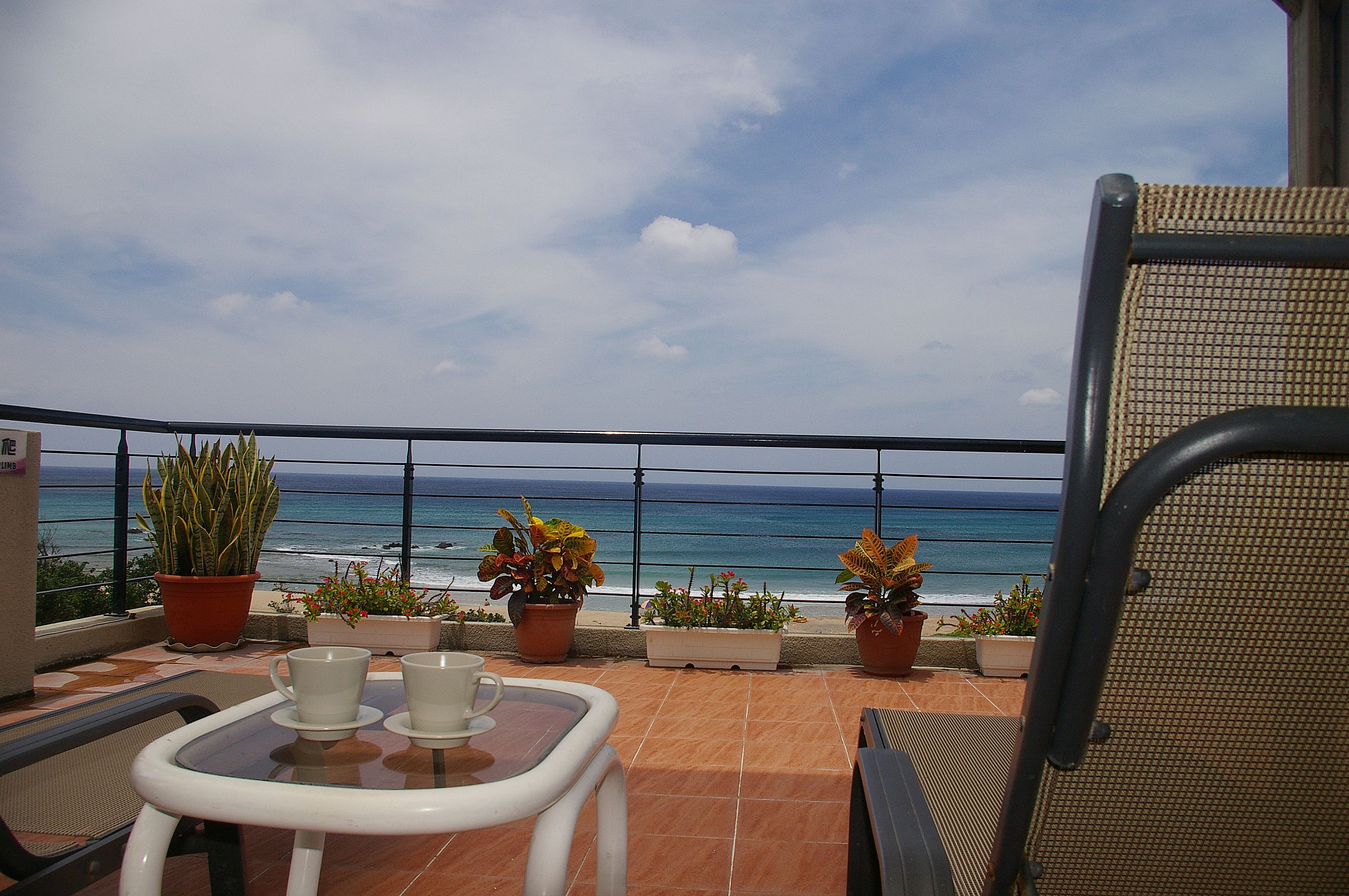 Un balcón con vistas al océano, plantas en macetas y una mesa con dos tazas.