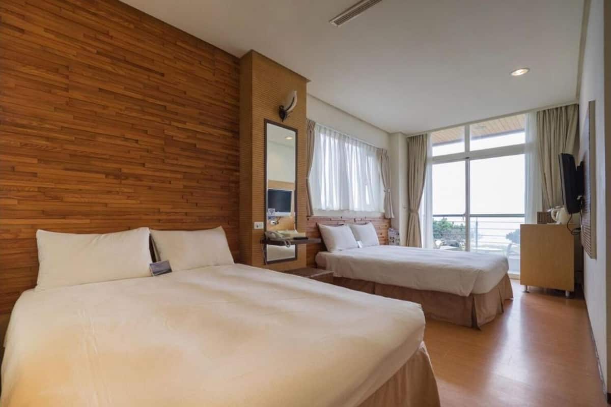 A hotel room with two beds, a wooden accent wall, a large window with curtains, and a television on a wooden cabinet.