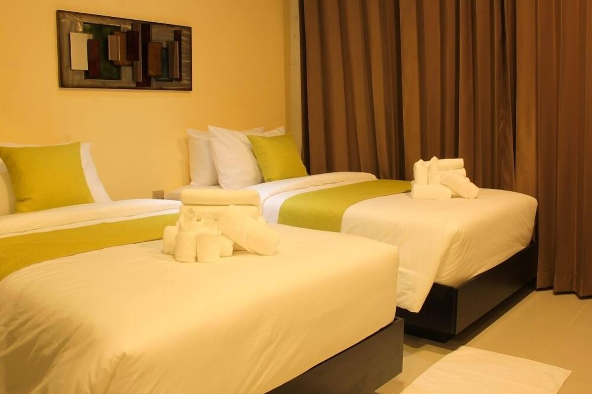 Ein Hotelzimmer mit zwei Betten, jeweils mit weißer Bettwäsche und grünen Akzenten, einem großen Fenster mit braunen Vorhängen und einem modernen, abstrakten Gemälde an der Wand.