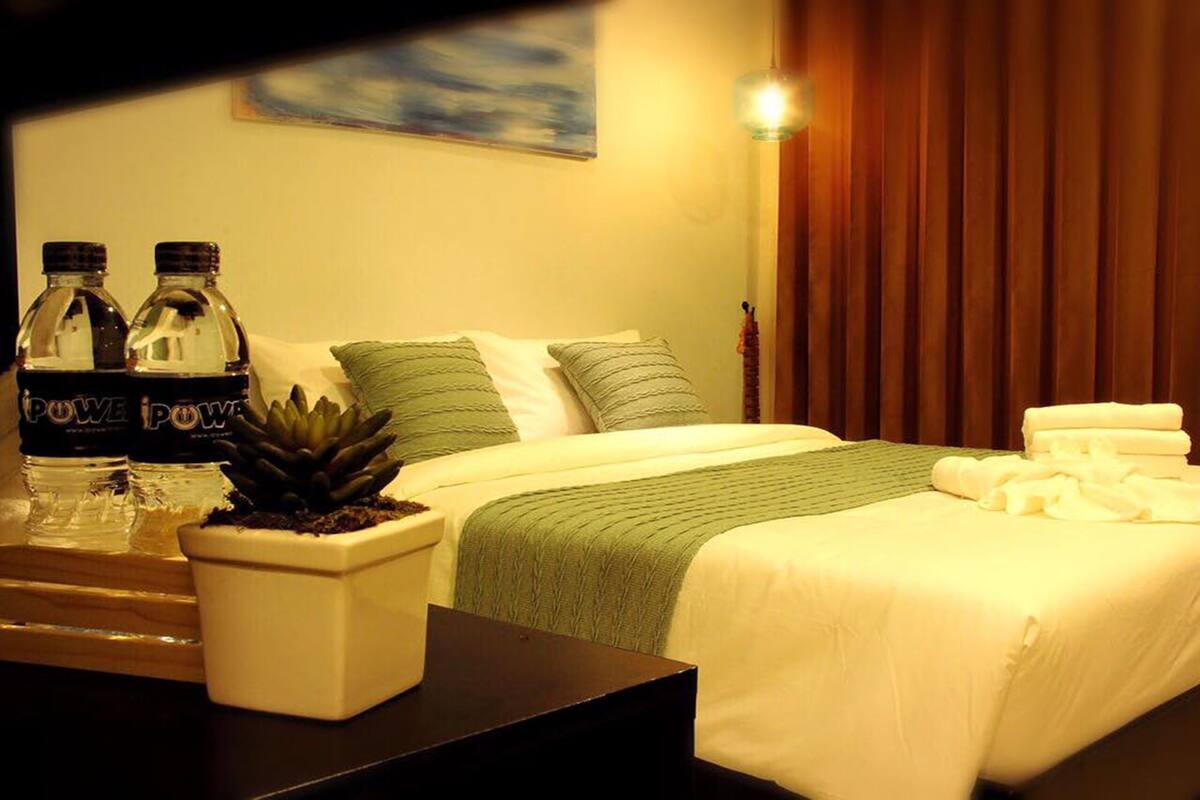 Ein Hotelzimmer mit Bett, Kissen, einem Nachttisch mit Wasserflaschen, einer Topfpflanze und einem Bild an der Wand.