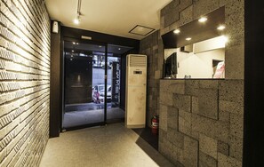 A modern hotel lobby with a brick wall, a glass door leading outside, a large air conditioning unit, and a fire extinguisher. - Blanc Hotel Gangnam (Seoul)