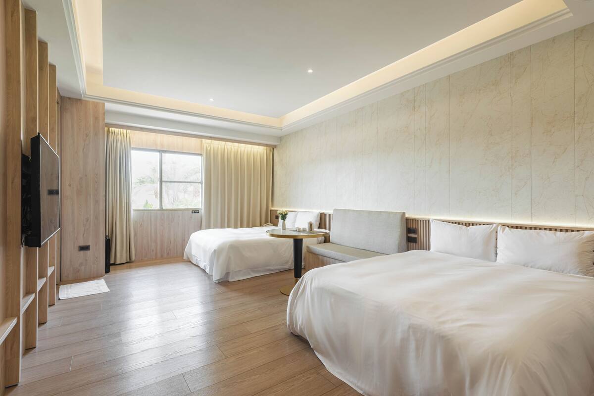 A hotel room with two beds, a TV, a round table, and a large window with curtains.
