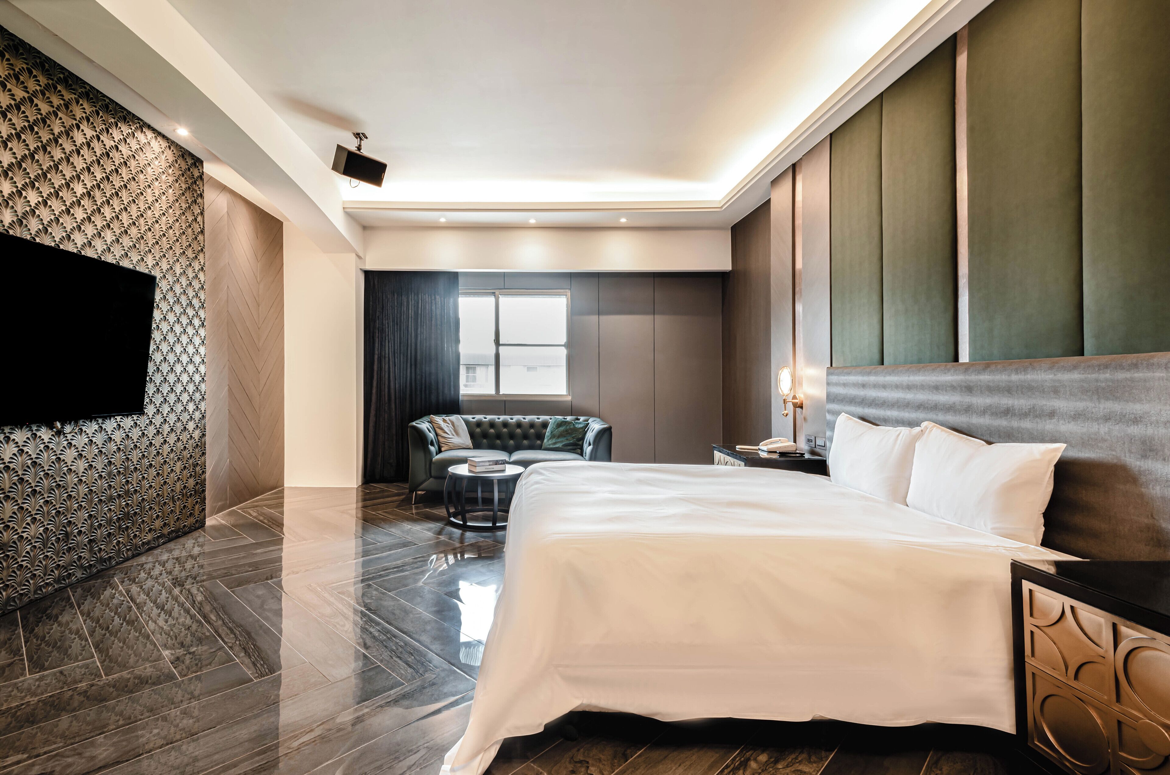 Una habitación de hotel moderna con una cama grande, un televisor de pantalla plana en la pared, una zona de estar con un sofá y una mesita redonda, y un ventanal que ofrece luz natural.
