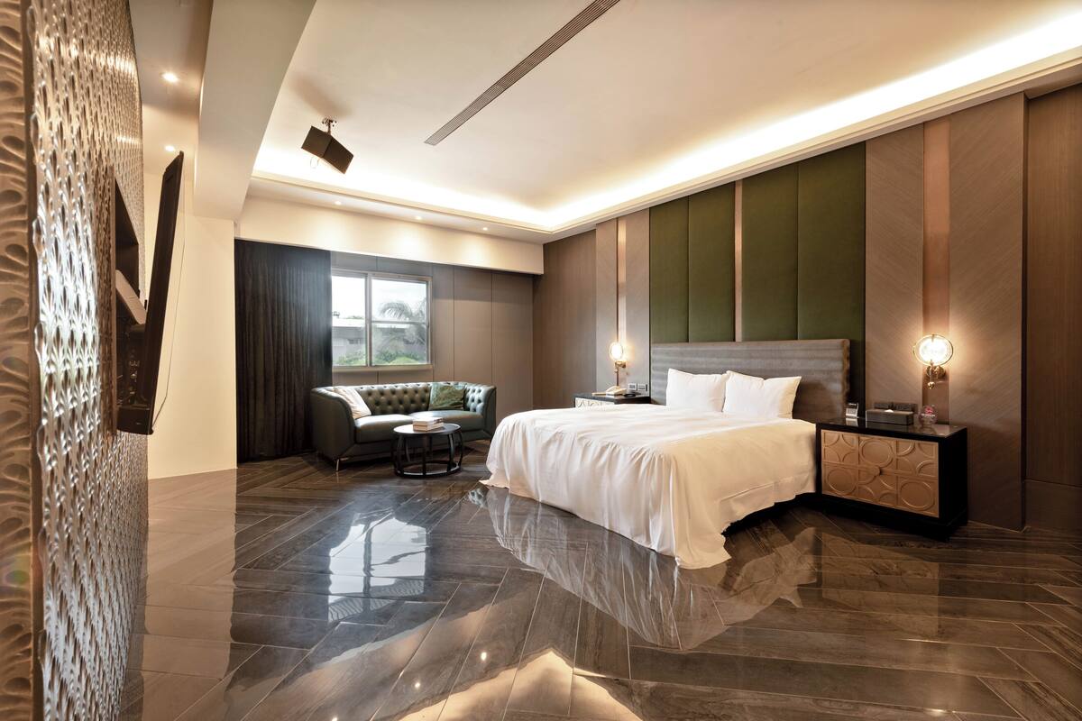 A modern hotel room with a large bed, a sofa, a small table, and a nightstand with a lamp.