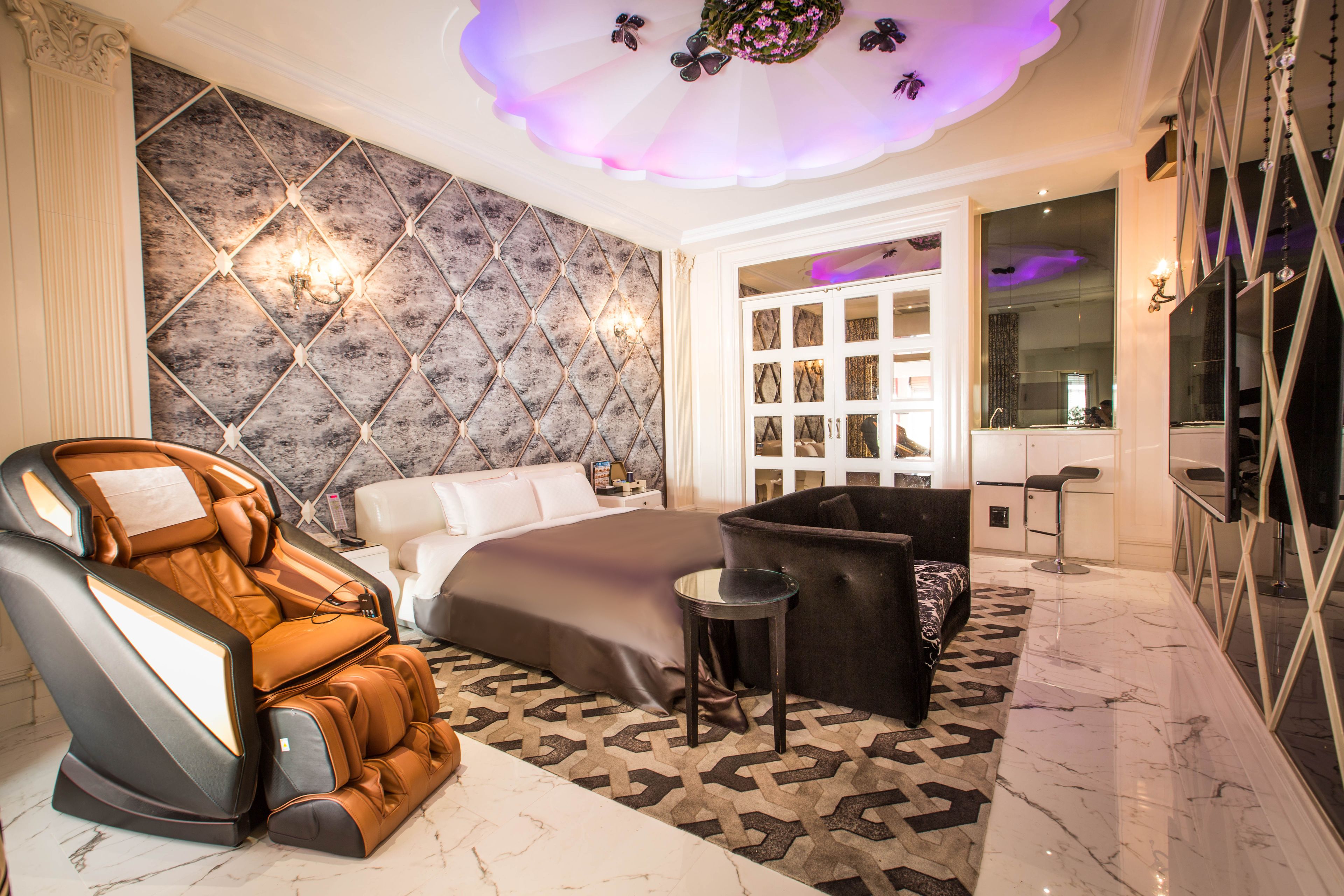 A modern bedroom with a massage chair, a large bed, a sofa, and a vanity with a mirror.