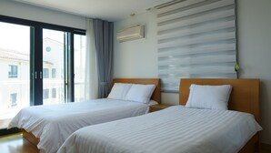 A hotel room with two single beds, a large window with curtains, a wall-mounted air conditioner, and a view of buildings outside. - Hera's Castle (Seogwipo)