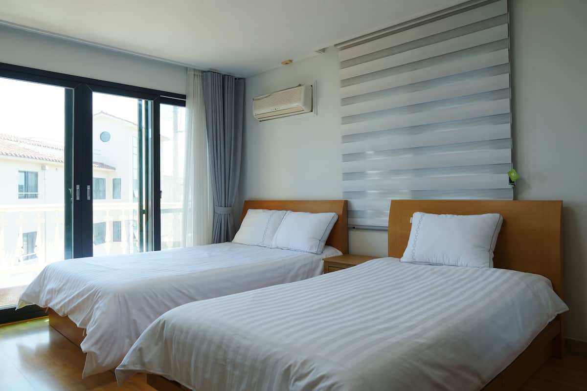Quarto de hotel com duas camas de solteiro, uma janela grande com cortinas, ar-condicionado fixado na parede e vista para prédios.
