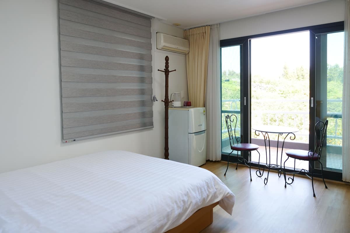 Quarto com cama, guarda-roupa, ar condicionado, varanda com mesa e cadeiras, e vista para o verde.