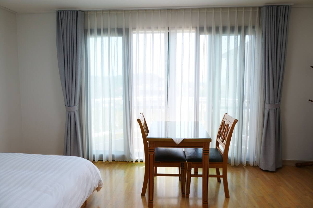 Quarto de hotel com cama, mesa de jantar e cadeiras, e janelas amplas com cortinas translúcidas.