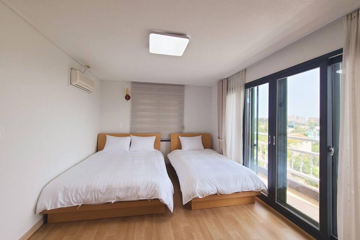 Quarto com duas camas, luminária no teto, aparelho de ar condicionado, janela com cortinas e vista para prédios.