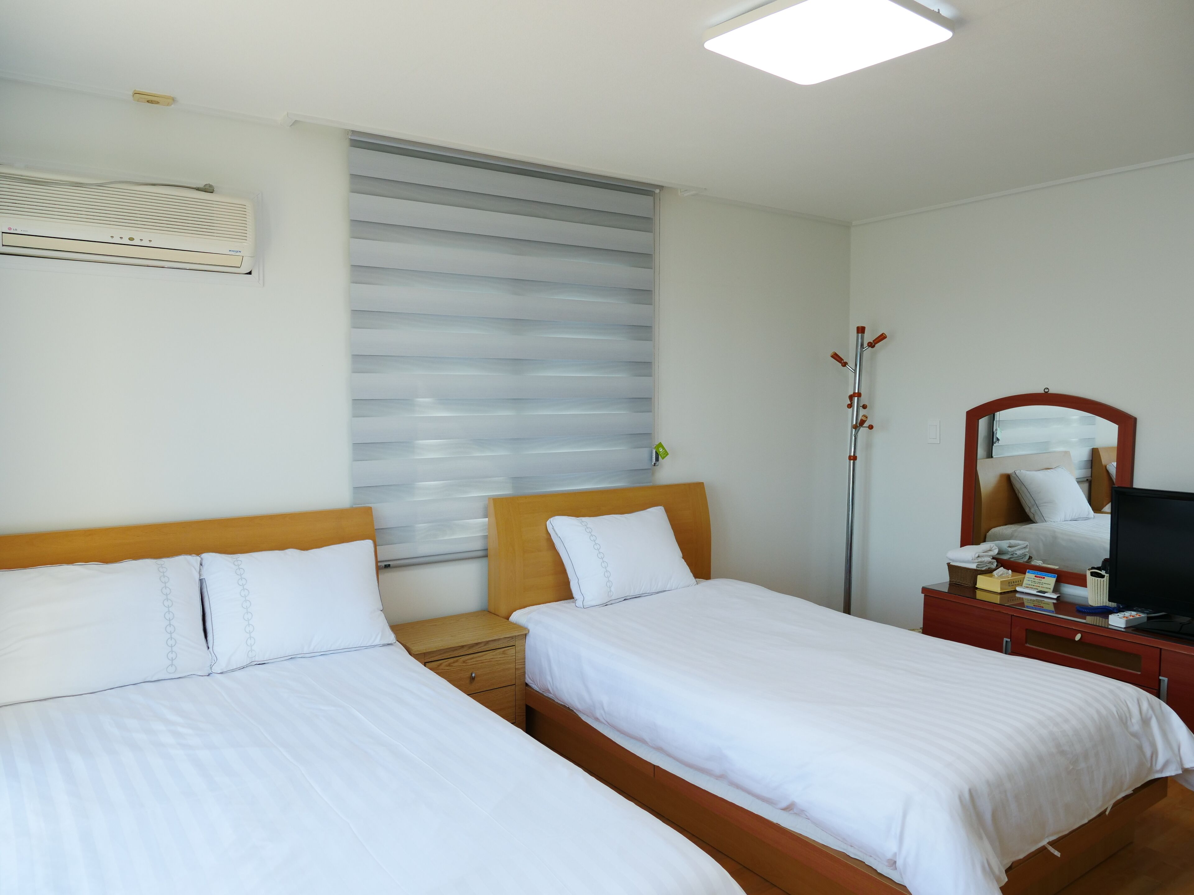 Habitación de hotel con dos camas individuales, una mesita de noche de madera, un espejo, un televisor y una ventana con persianas.
