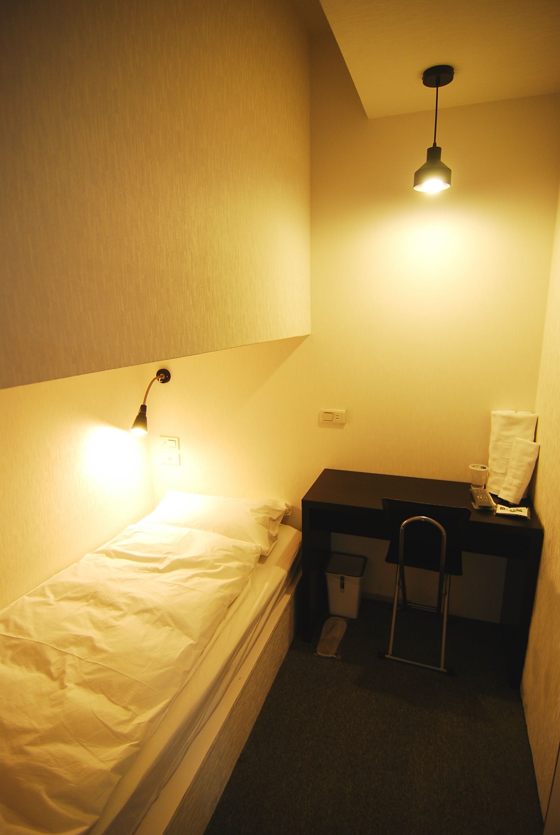 A small, dimly lit room with a single bed, a desk, and a hanging light fixture.