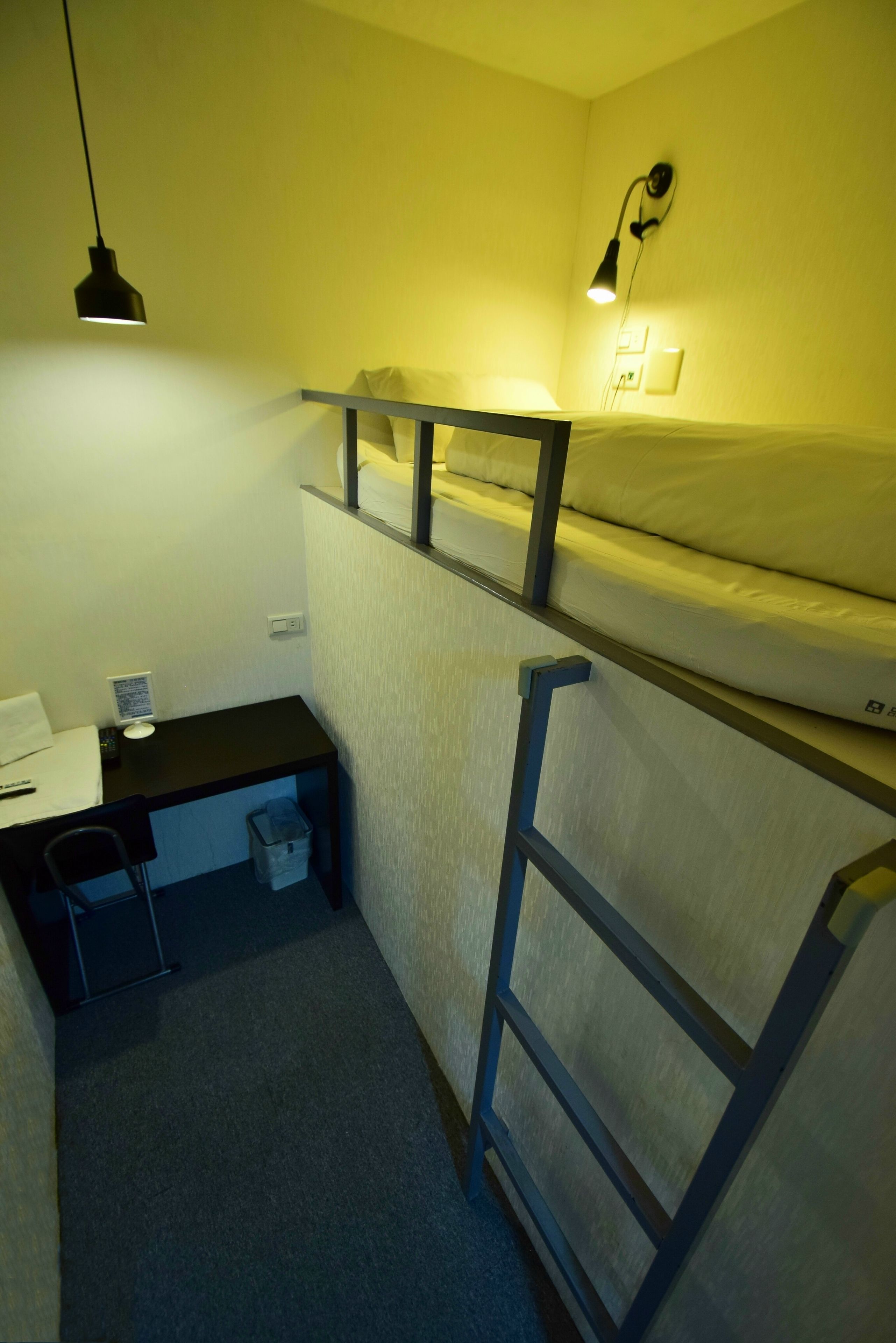 A compact hotel room with a bunk bed, a desk with a chair, a small trash bin, and a wall-mounted lamp.