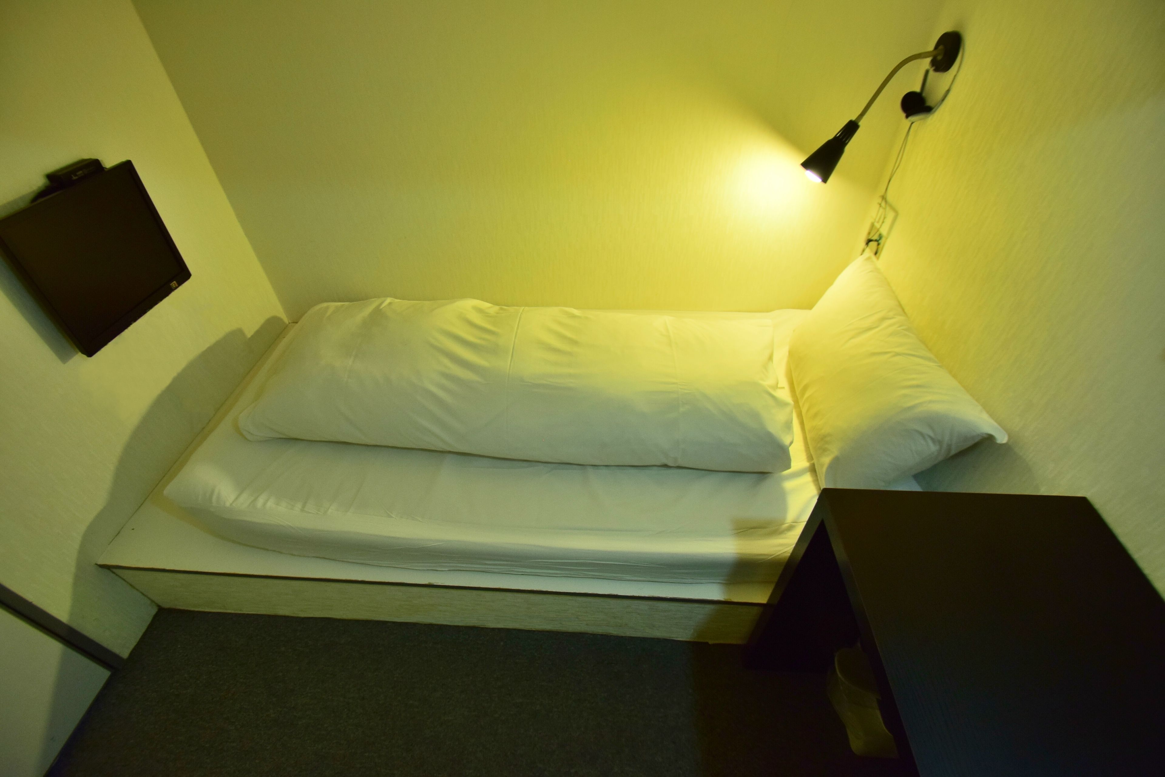 A small, single-bed room with a bedside table, a wall-mounted lamp, and a flat-screen TV on the wall.