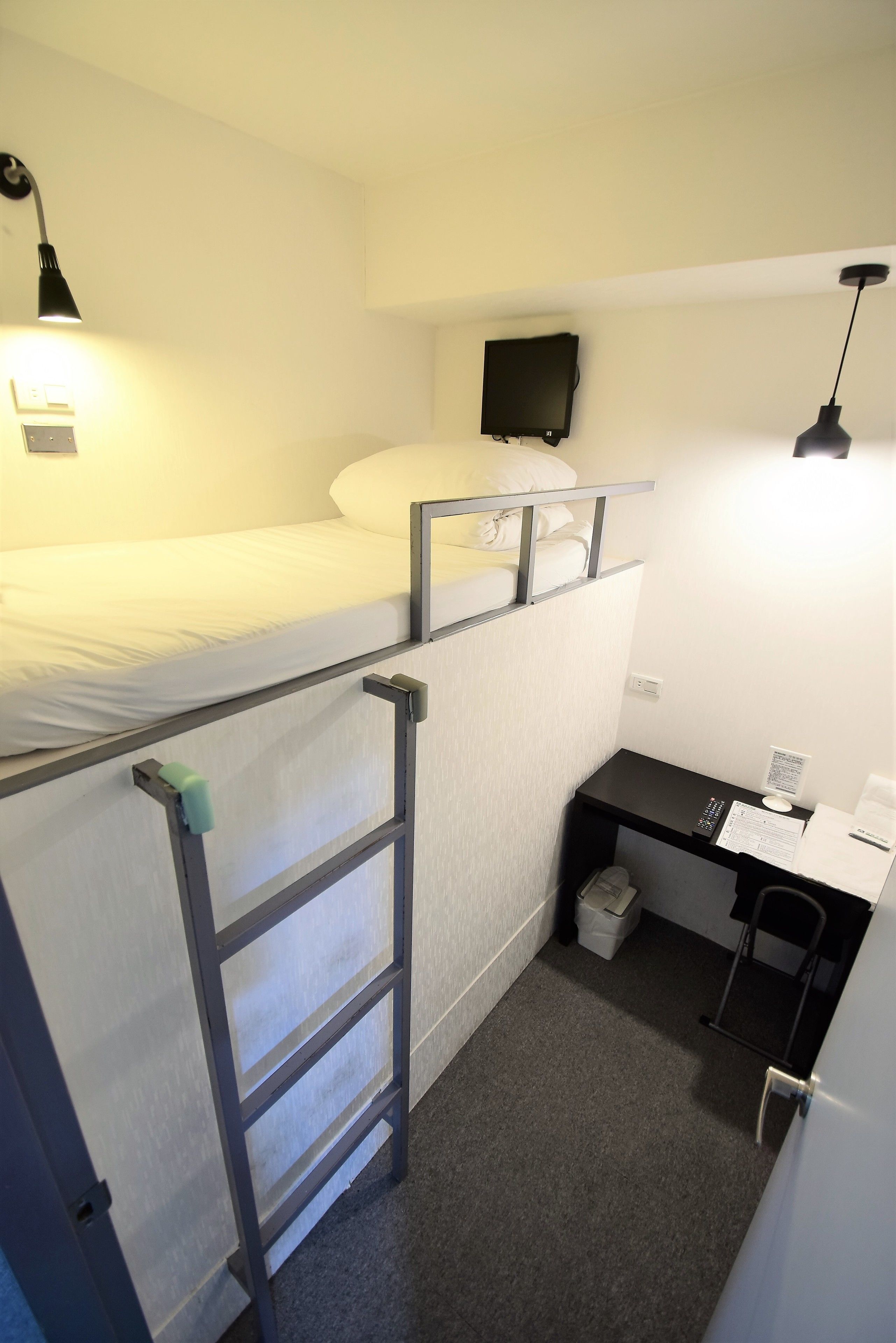 Een stapelbed met een bureau en stoel, een kleine prullenbak en een aan de muur bevestigde televisie.